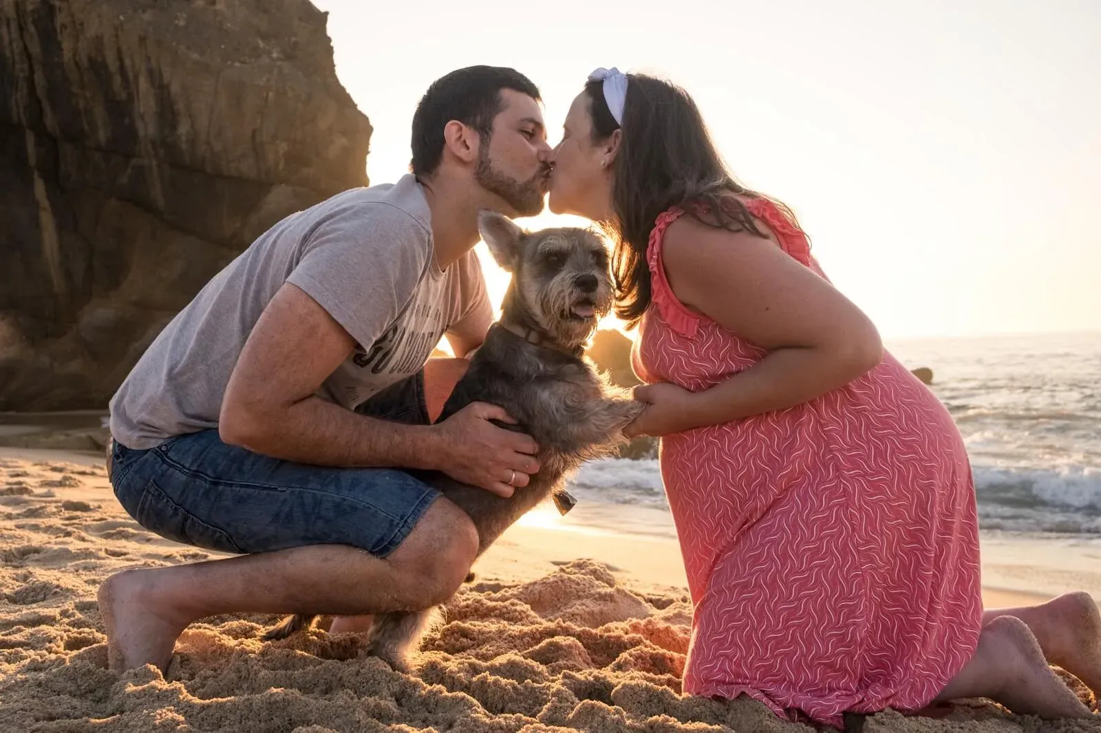 Casal ajoelhado na areia a beijar-se, com o cão